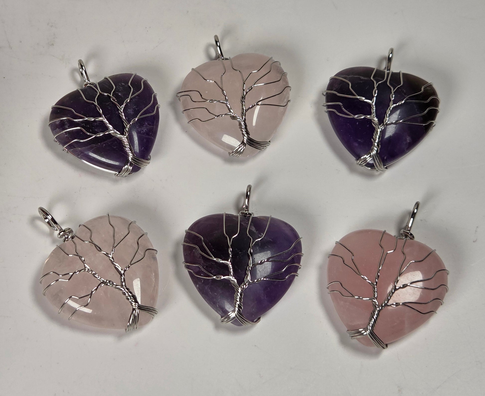 Six heart-shaped crystal pendants arranged in two rows on a light gray background. The pendants feature tree-like silver wire patterns set into polished stones in shades of purple, lavender, and pale pink.