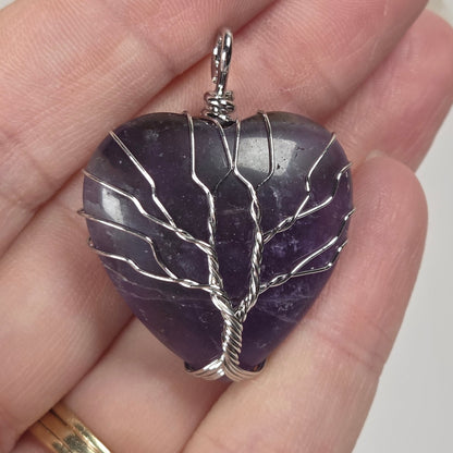 A teardrop-shaped purple stone pendant wrapped in silver wire, held in a person's hand. The pendant features a polished stone with silver wire branches forming a leaf or tree-like pattern.