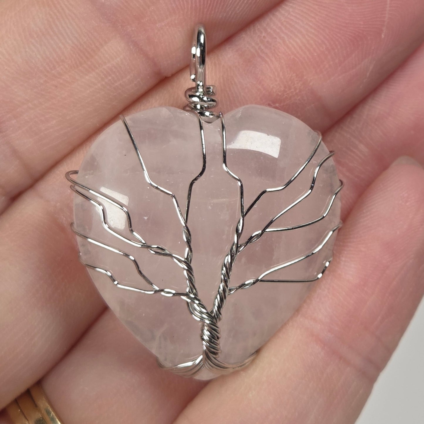A close-up of a hand holding a clear, leaf-shaped stone pendant with silver wire wrapped in a tree-like pattern. The hand is shown against a plain light background, and a gold ring is visible on the finger.