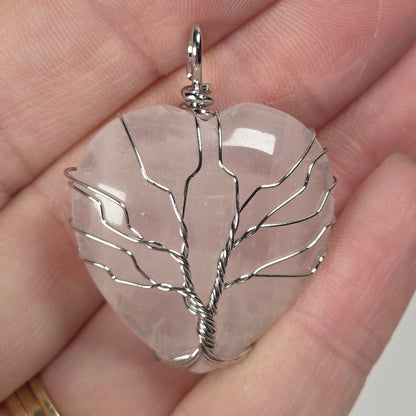A close-up of a hand holding a clear, leaf-shaped stone pendant with silver wire wrapped in a tree-like pattern. The hand is shown against a plain light background, and a gold ring is visible on the finger.