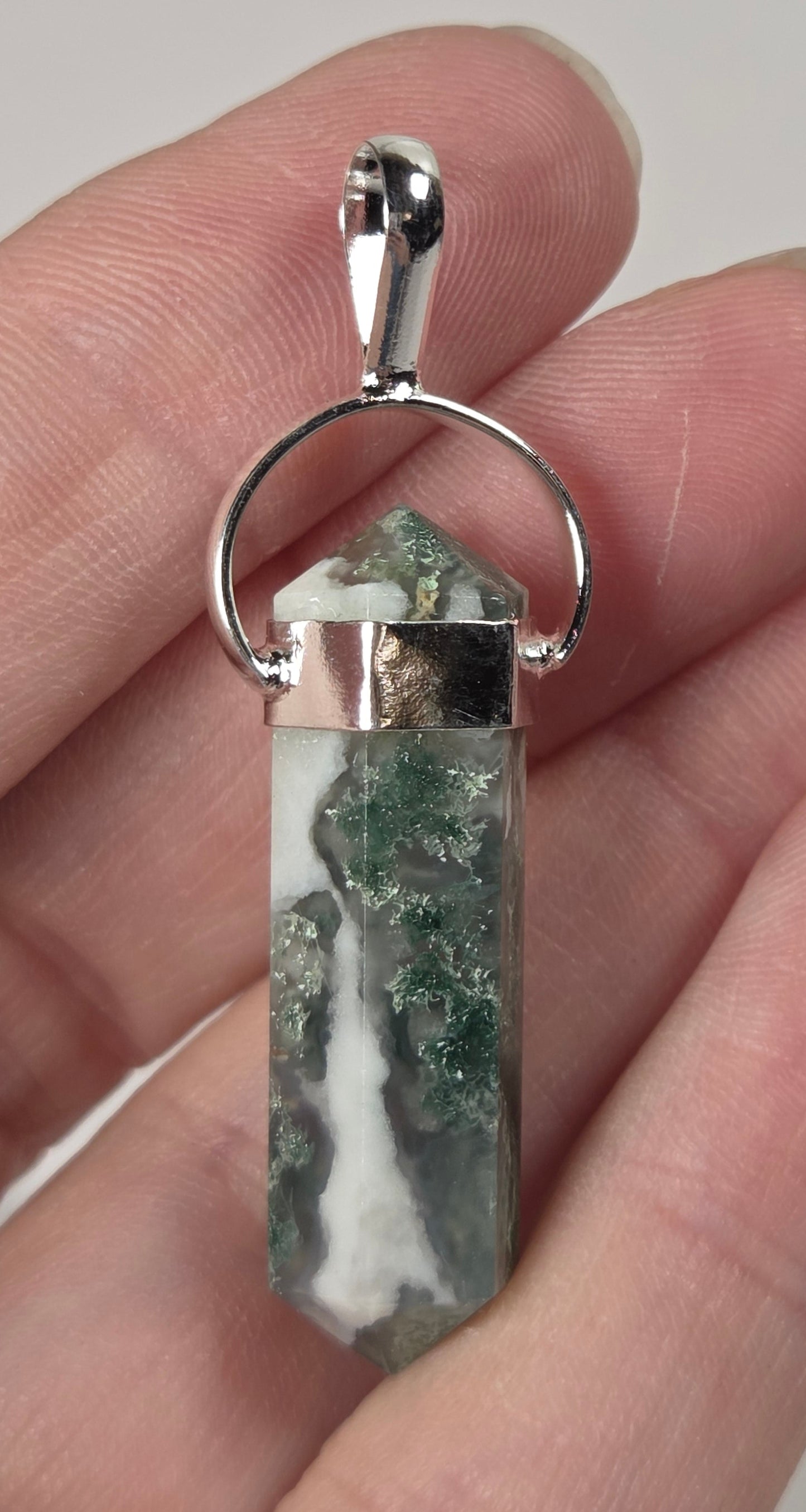 A silver-toned metal pendant with a polished green and white marbled crystal point, held in a person's hand against a plain light background.