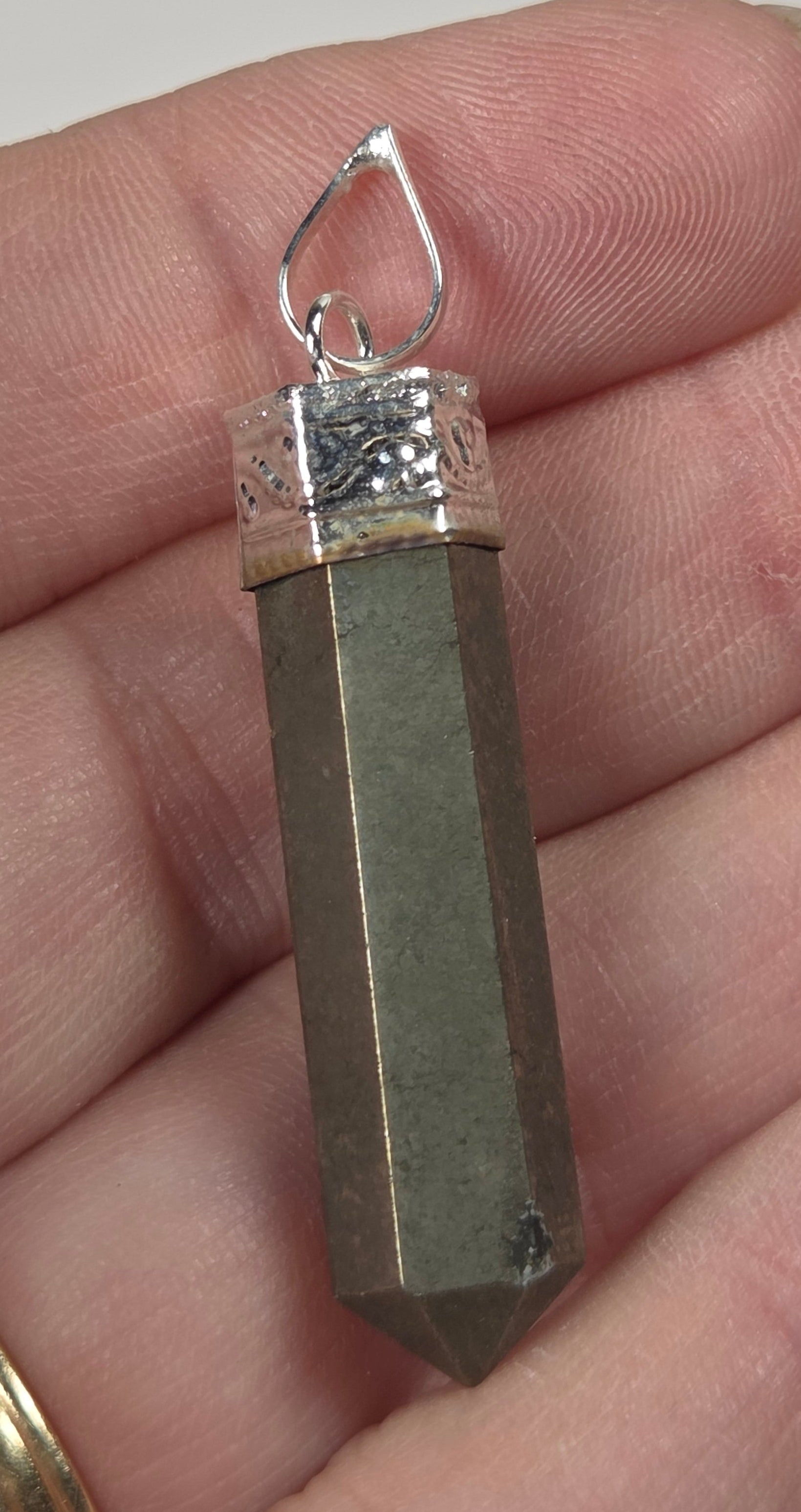A silver-toned metal pendant shaped like a pointed crystal tower, held in a person's hand. The pendant features a silver bail and a textured silver band at the top, with a polished metallic gray body.