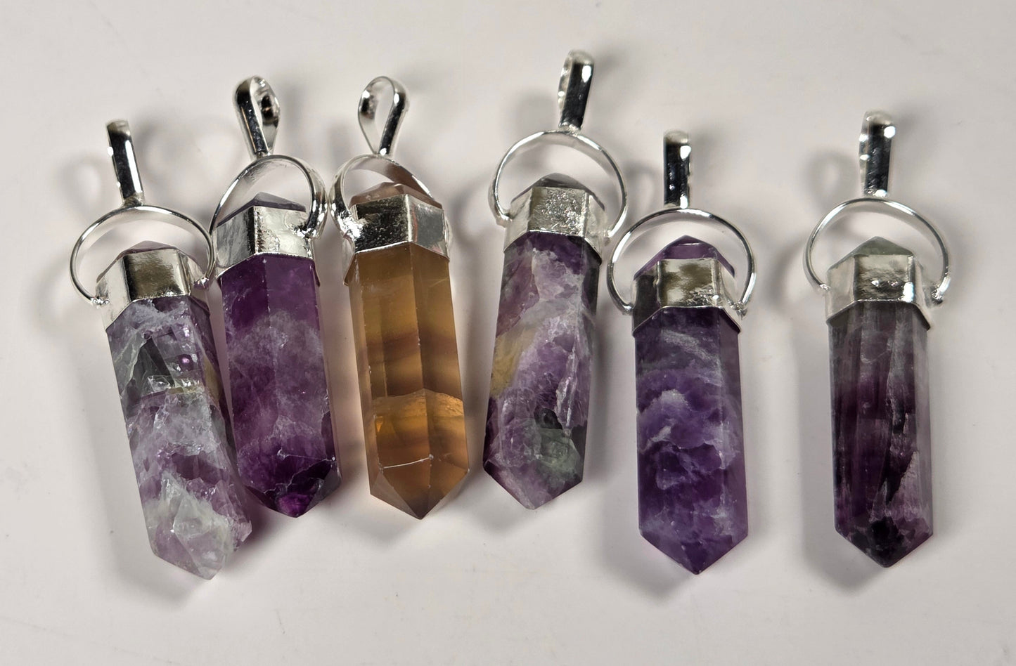 Five raw crystal point pendants with silver metal caps and loops, displayed on a light off-white background. The crystals feature natural marbling in shades of purple, brown, and gray.