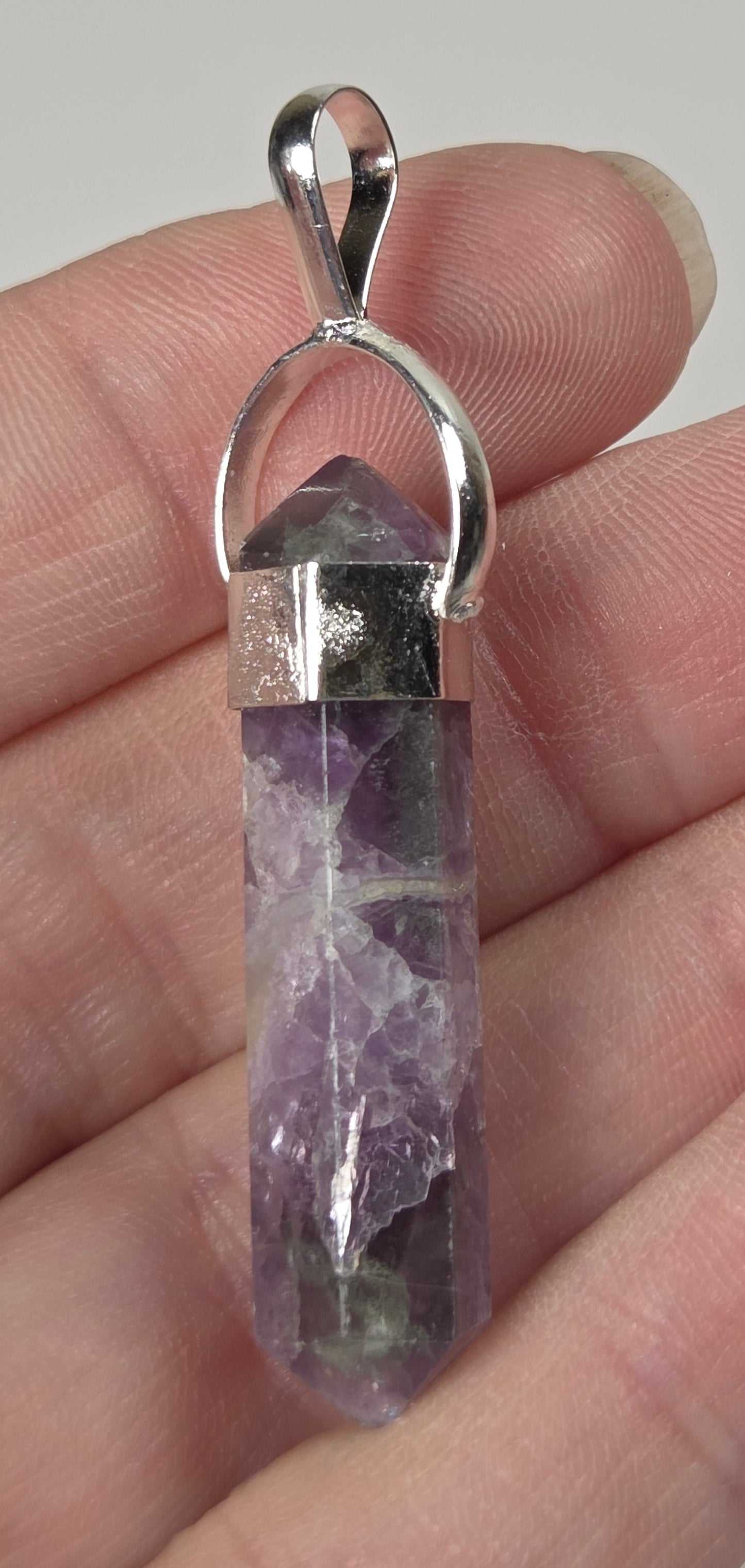 A silver pendant featuring a natural purple amethyst crystal point, held in a person's hand against a neutral light gray background.