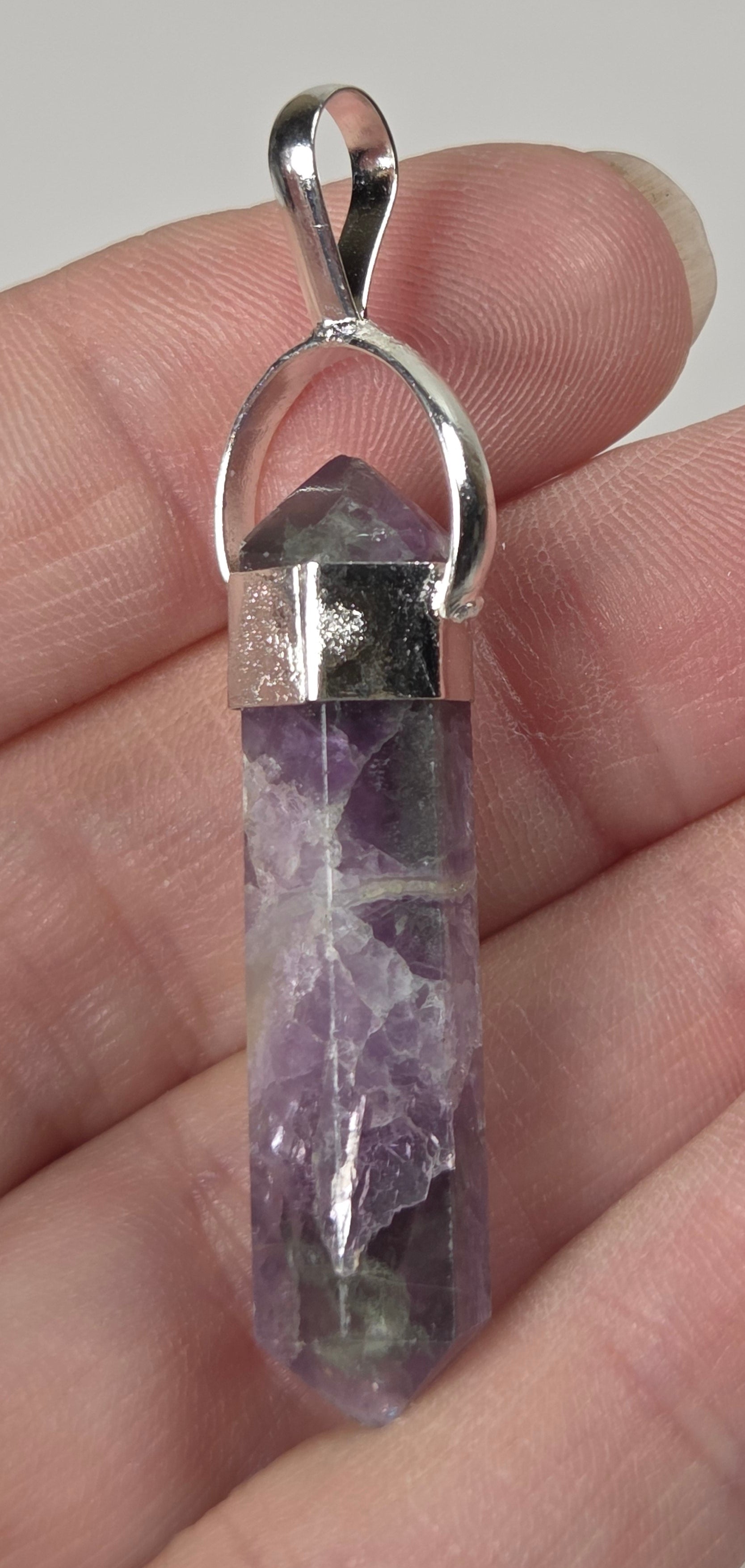 A silver pendant featuring a natural purple amethyst crystal point, held in a person's hand against a neutral light gray background.