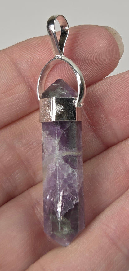A silver pendant featuring a natural purple amethyst crystal point, held in a person's hand against a neutral light gray background.