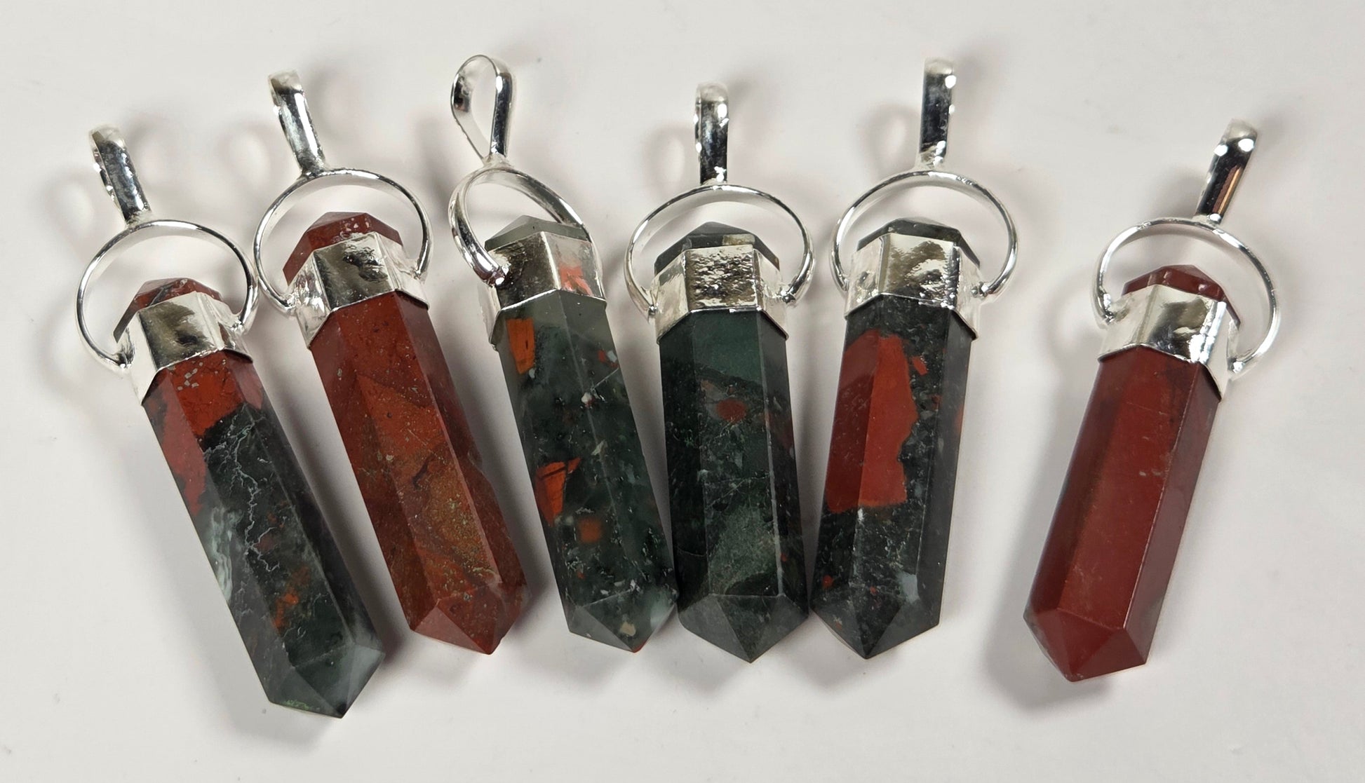 Six faceted crystal pendants with silver-toned metal caps and loops, arranged vertically on a plain white background. The crystals alternate between dark green with red inclusions and reddish-brown with dark inclusions.