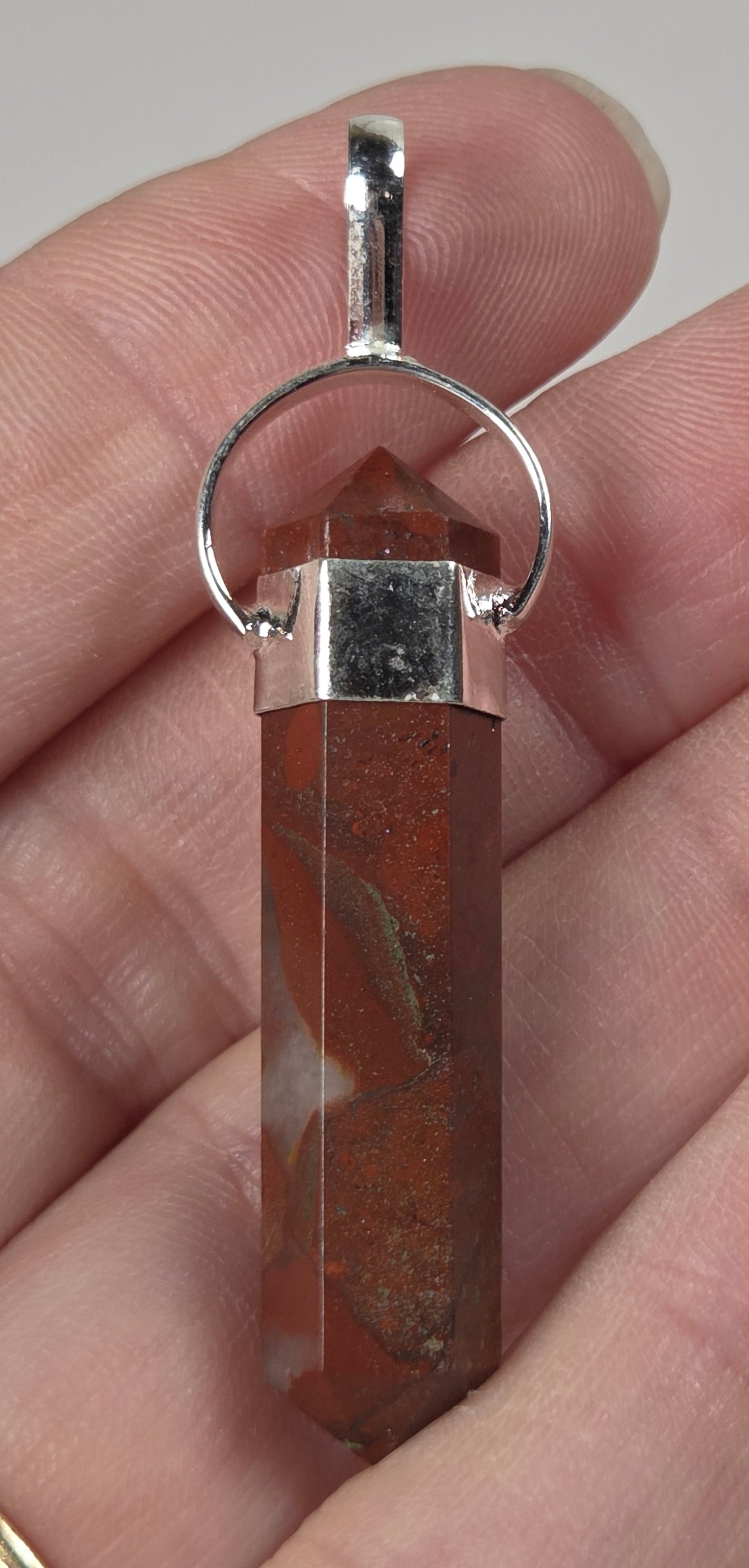 A silver-toned cylindrical crystal pendant with a natural reddish-brown agate pattern, held in a person's hand.