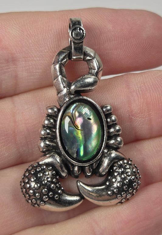A silver lobster claw pendant with an iridescent abalone shell center, held on a person's fingers against a light gray background.