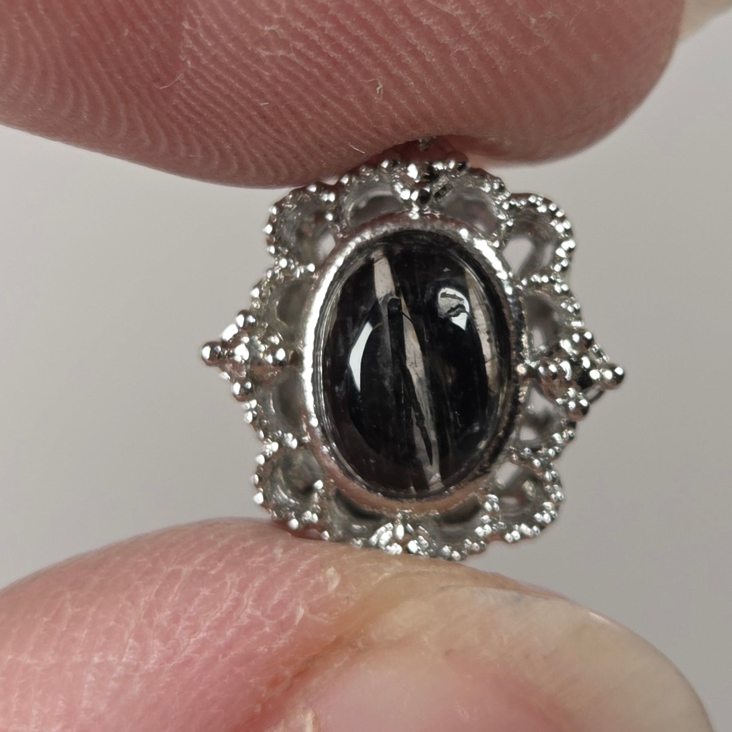 A close-up of a silver-toned ring with a black oval center stone and small clear stones around it, held between two fingers against a light gray background.