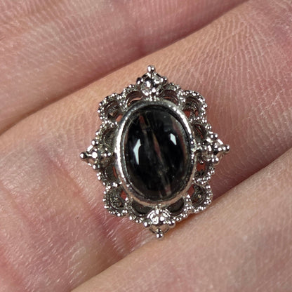 A silver-toned ring with an oval black cabochon stone and small clear stones around it, shown worn on a finger.