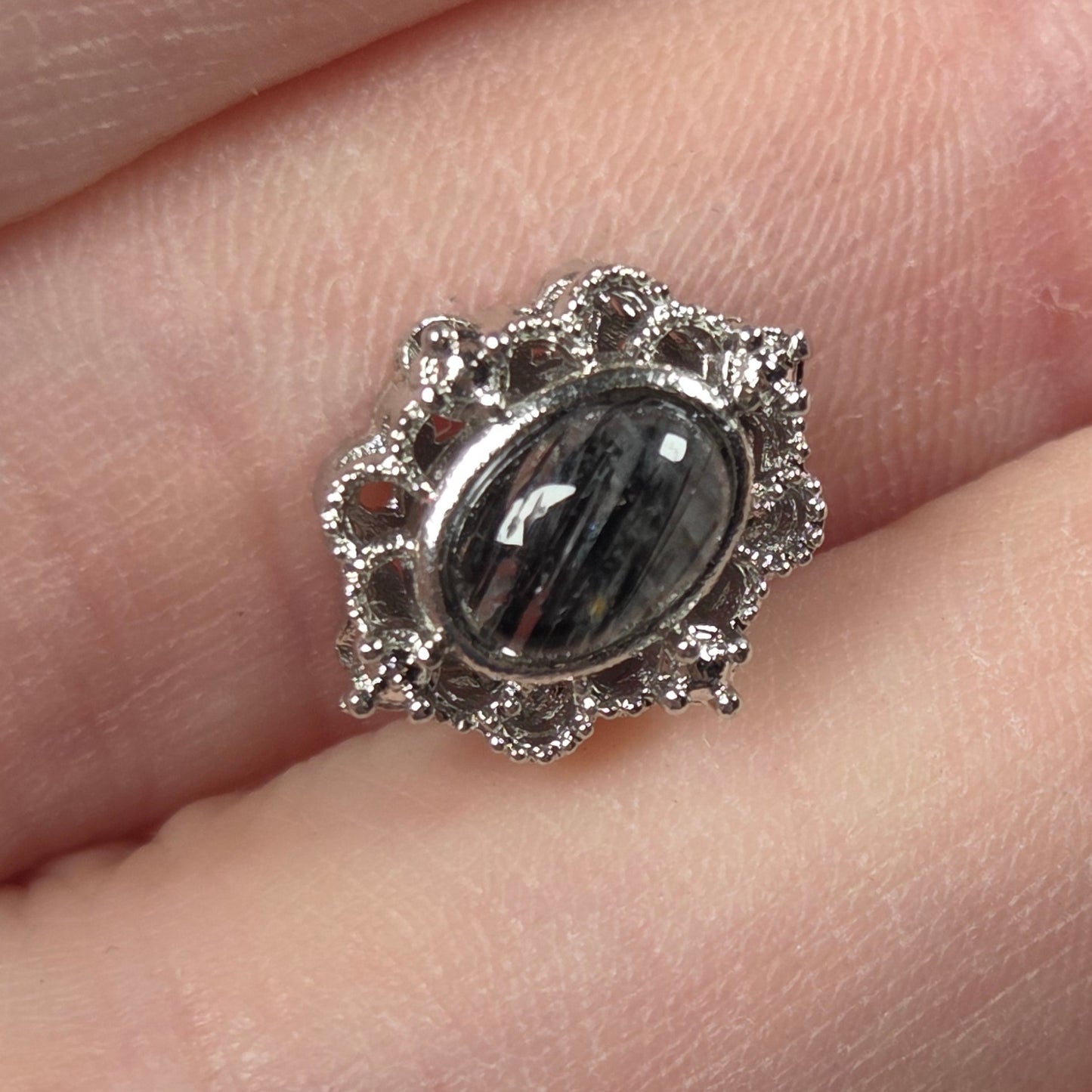 A silver-toned ring featuring an oval black and gray banded gemstone, set in a decorative scalloped halo with small clear stones, shown worn on a finger.
