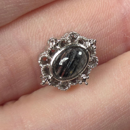 A silver-toned ring featuring an oval black and gray banded gemstone, set in a decorative scalloped halo with small clear stones, shown worn on a finger.