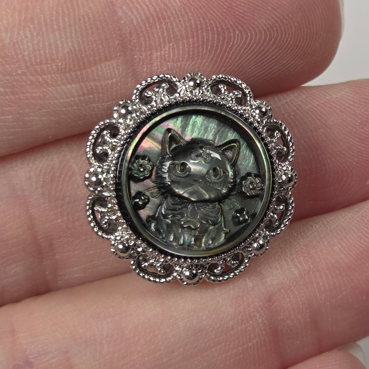 A close-up of a hand holding a round silver-toned charm featuring a raised cat face and floral filigree border, with an iridescent green-gray center.