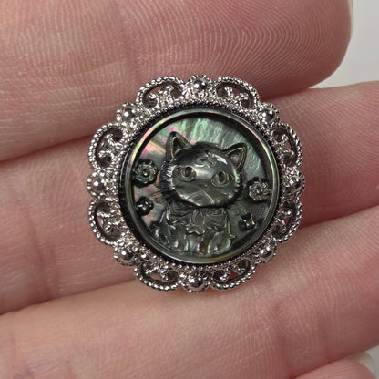A close-up of a hand holding a round silver-toned charm featuring a raised cat face and floral filigree border, with an iridescent green-gray center.