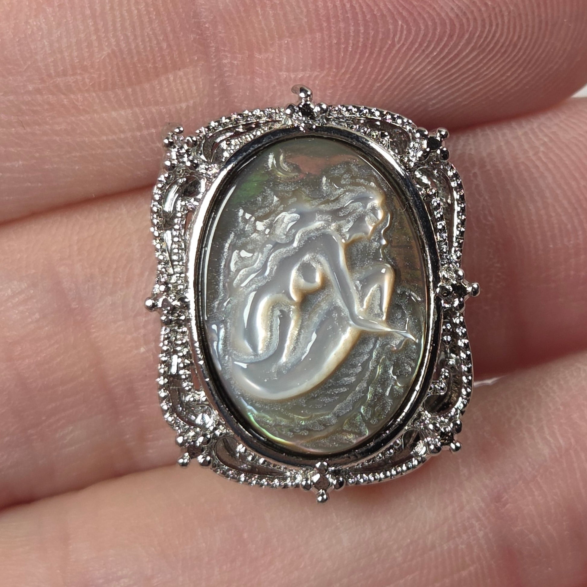 A silver-toned ring with an oval carved cabochon featuring a mermaid or woman figure, displayed on a person's fingers.