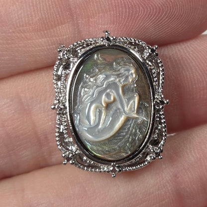 A silver-toned ring with an oval carved cabochon featuring a mermaid or woman figure, displayed on a person's fingers.