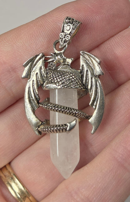 A silver-toned dragon pendant holding a clear quartz crystal point, held in a person's hand against a neutral light gray background.