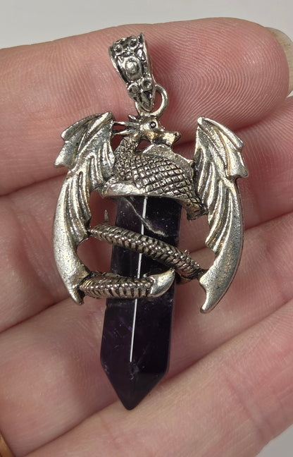 A silver-toned dragon pendant holding a dark purple crystal point, held in a person's hand against a plain light background.