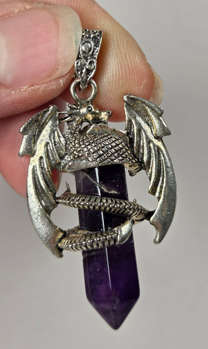 A hand holding a silver dragon pendant wrapped around a purple amethyst crystal point, shown against a plain light gray background.