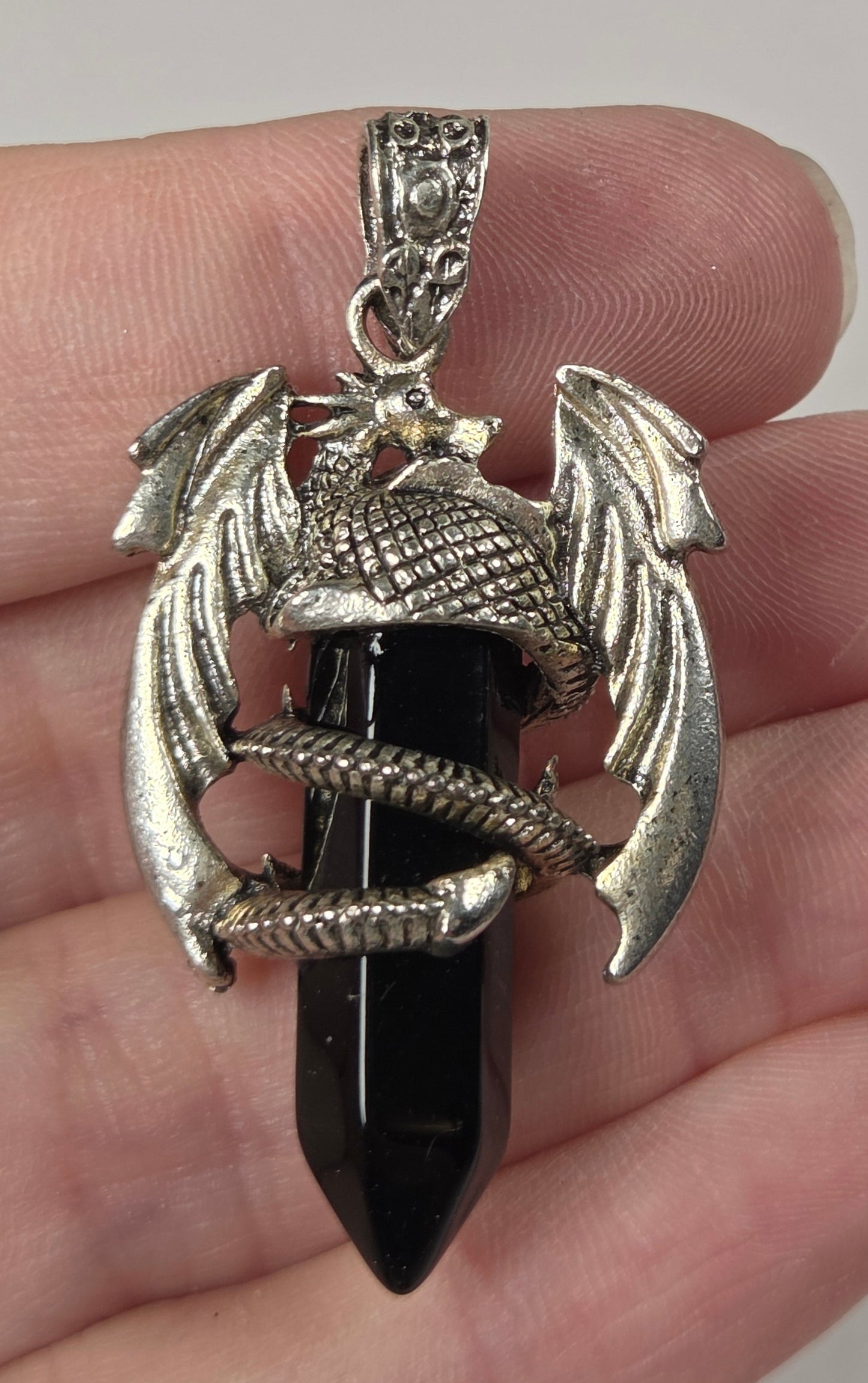 A silver-toned dragon pendant wrapped around a black pointed crystal, held in a person's hand against a plain light background.