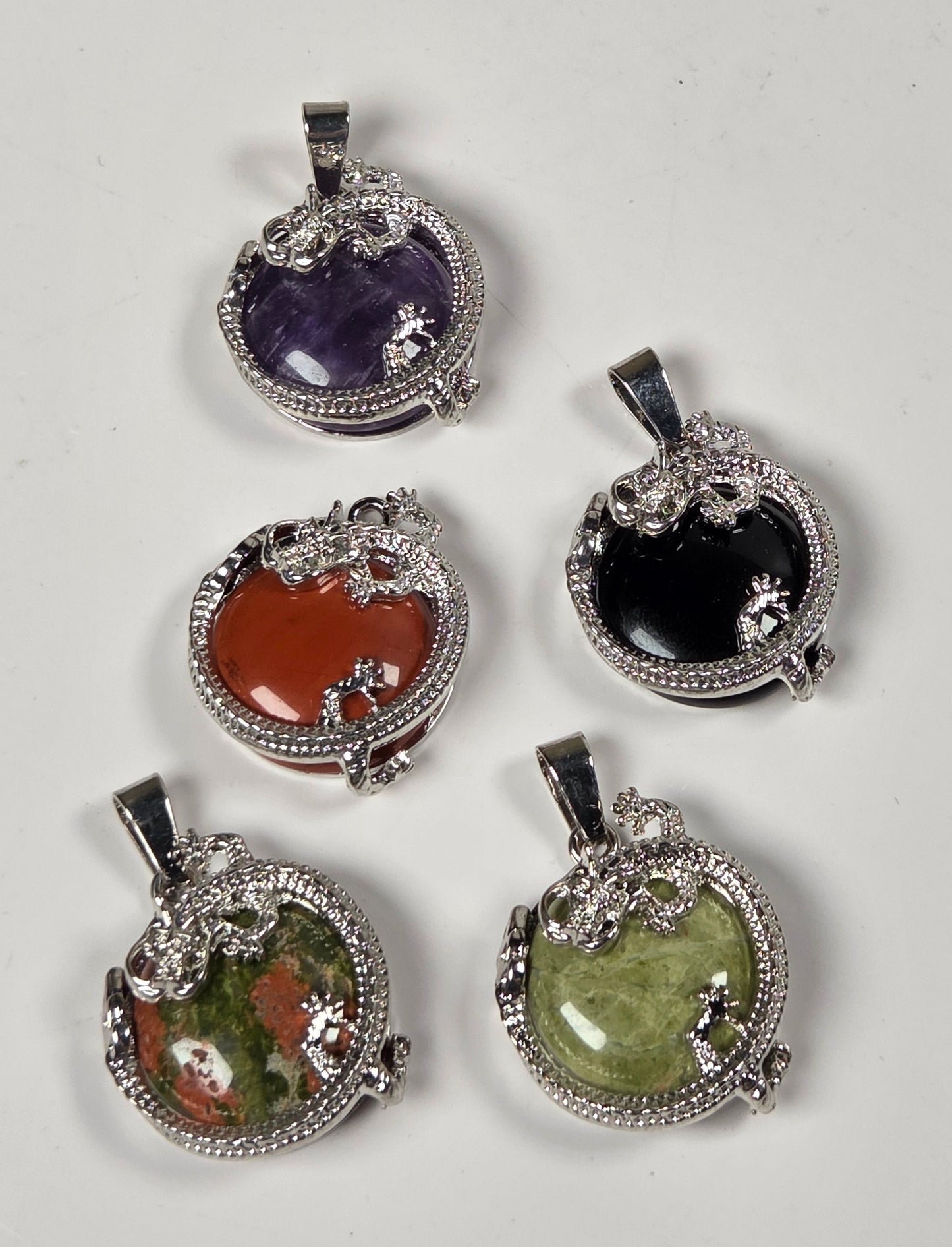 Five oval gemstone pendants with silver-toned milgrain-style borders and bail loops, arranged on a plain light background. The stones include black, green, purple, orange, and a mottled green-and-brown stone.