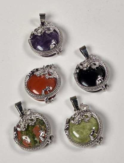 Five oval gemstone pendants with silver-toned milgrain-style borders and bail loops, arranged on a plain light background. The stones include black, green, purple, orange, and a mottled green-and-brown stone.