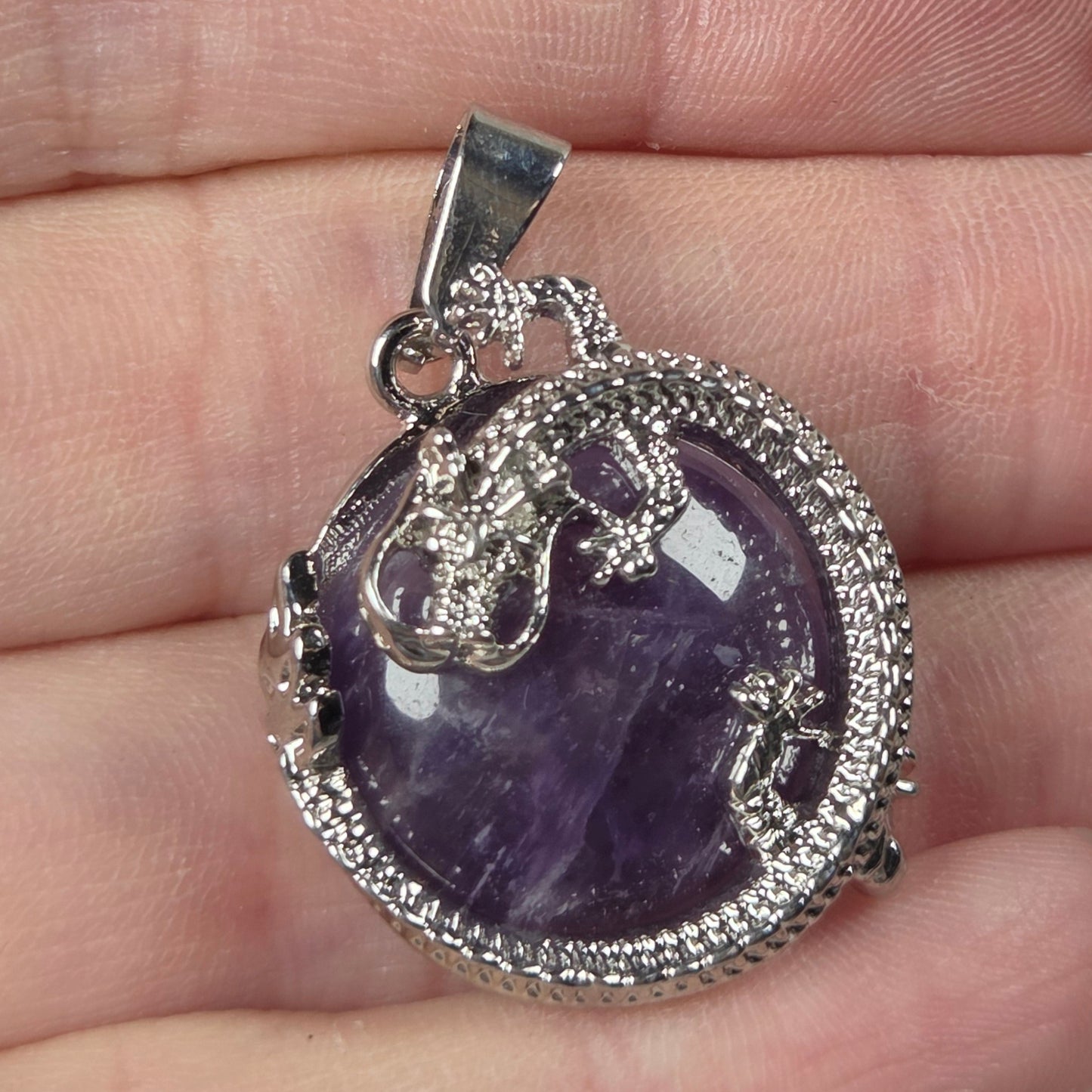 A silver-toned pendant with a purple amethyst cabochon and pavé-set clear stones, held on a person's fingers against a light gray background.