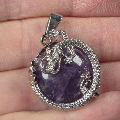A silver-toned pendant with a purple amethyst cabochon and pavé-set clear stones, held on a person's fingers against a light gray background.