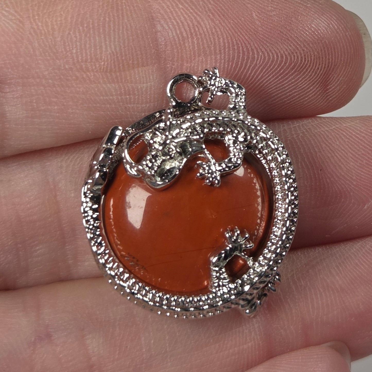 A silver-toned pendant with a polished orange-brown heart-shaped center stone, held on a person's fingers against a plain light background.