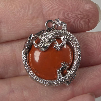 A silver-toned pendant with a polished orange-brown heart-shaped center stone, held on a person's fingers against a plain light background.