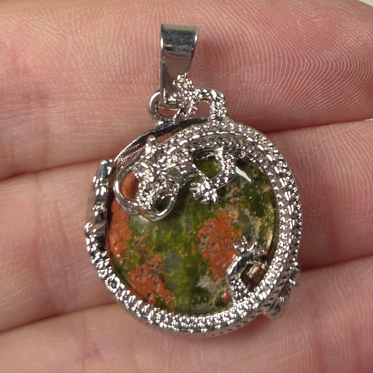 A silver-toned pendant with a textured green and orange stone center, held on a person's fingers against a plain light background.