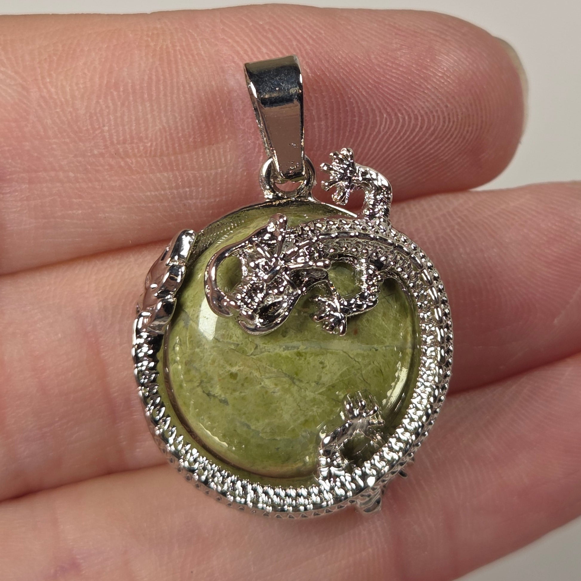 A silver pendant with a green heart-shaped stone and a silver dragon design, resting on a person's fingers against a light beige background.
