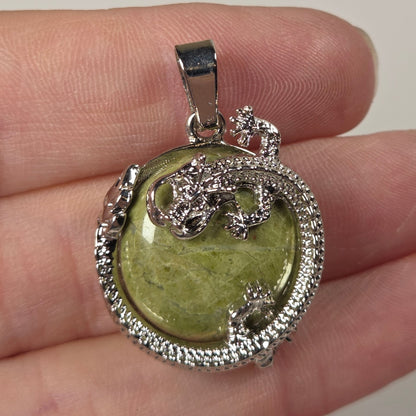 A silver pendant with a green heart-shaped stone and a silver dragon design, resting on a person's fingers against a light beige background.