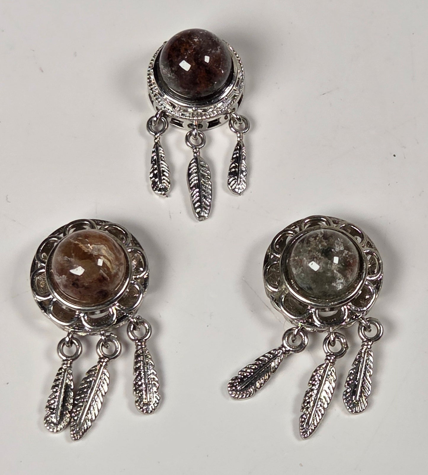 Three silver-toned dangle earrings with oval gemstone settings and feather-like dangling charms, arranged on a plain light background.