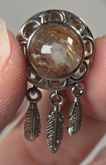A close-up of a hand holding a silver-toned pendant featuring a round brownish gemstone and three dangling feather charms.