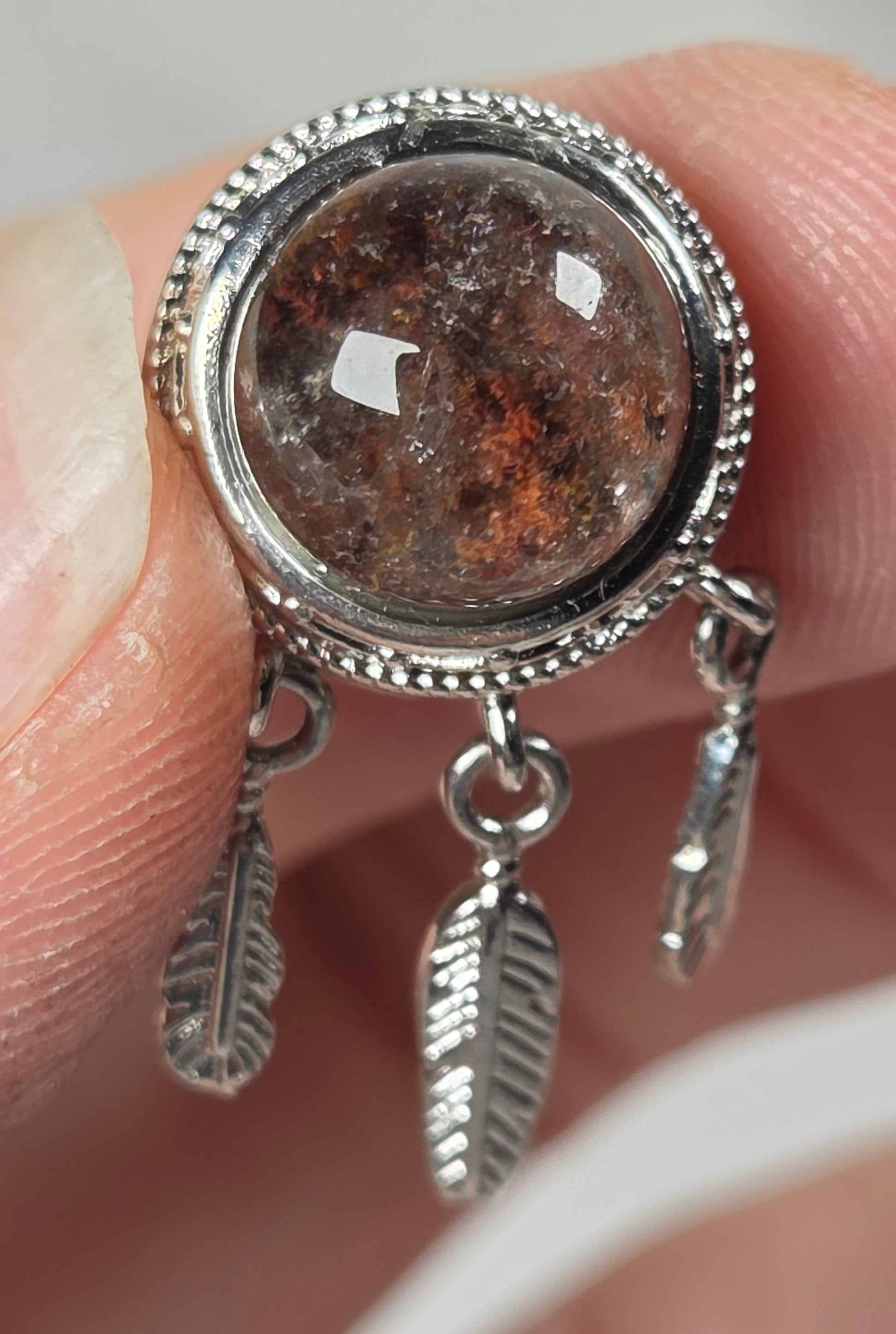 A close-up of a silver-toned ring held between fingers, featuring a round brown textured stone in a beaded silver bezel with two small feather-shaped dangling charms.