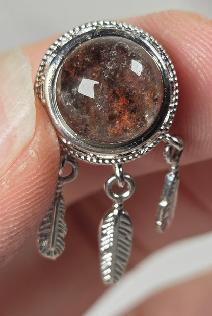A close-up of a silver-toned ring held between fingers, featuring a round brown textured stone in a beaded silver bezel with two small feather-shaped dangling charms.
