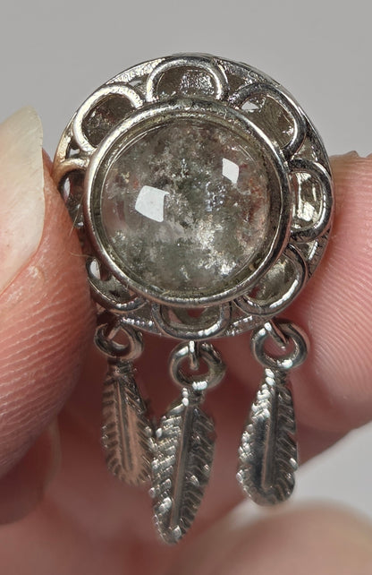 A close-up of a hand holding a silver-toned ring with a round textured stone and two dangling feather-shaped charms.