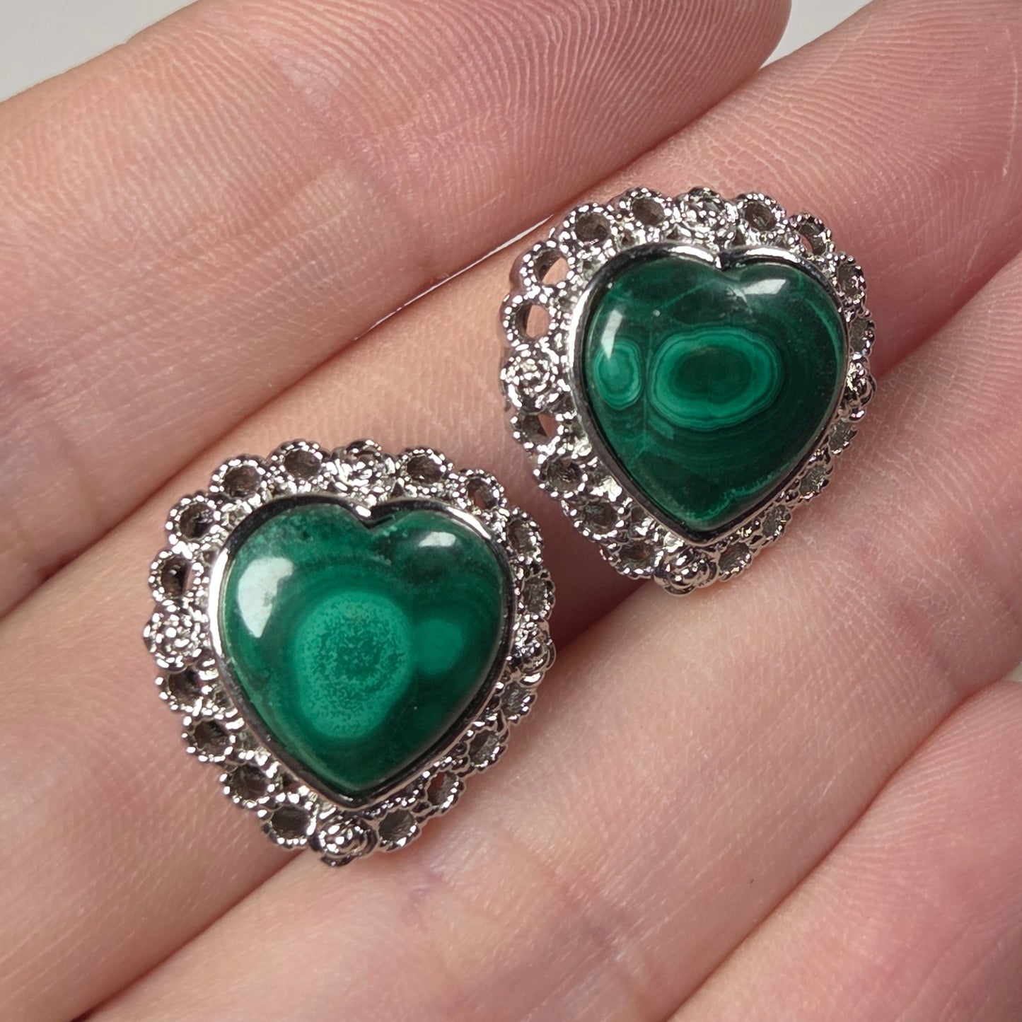 A pair of heart-shaped green malachite stud earrings with silver settings, shown resting on a person's fingers against a light gray background.