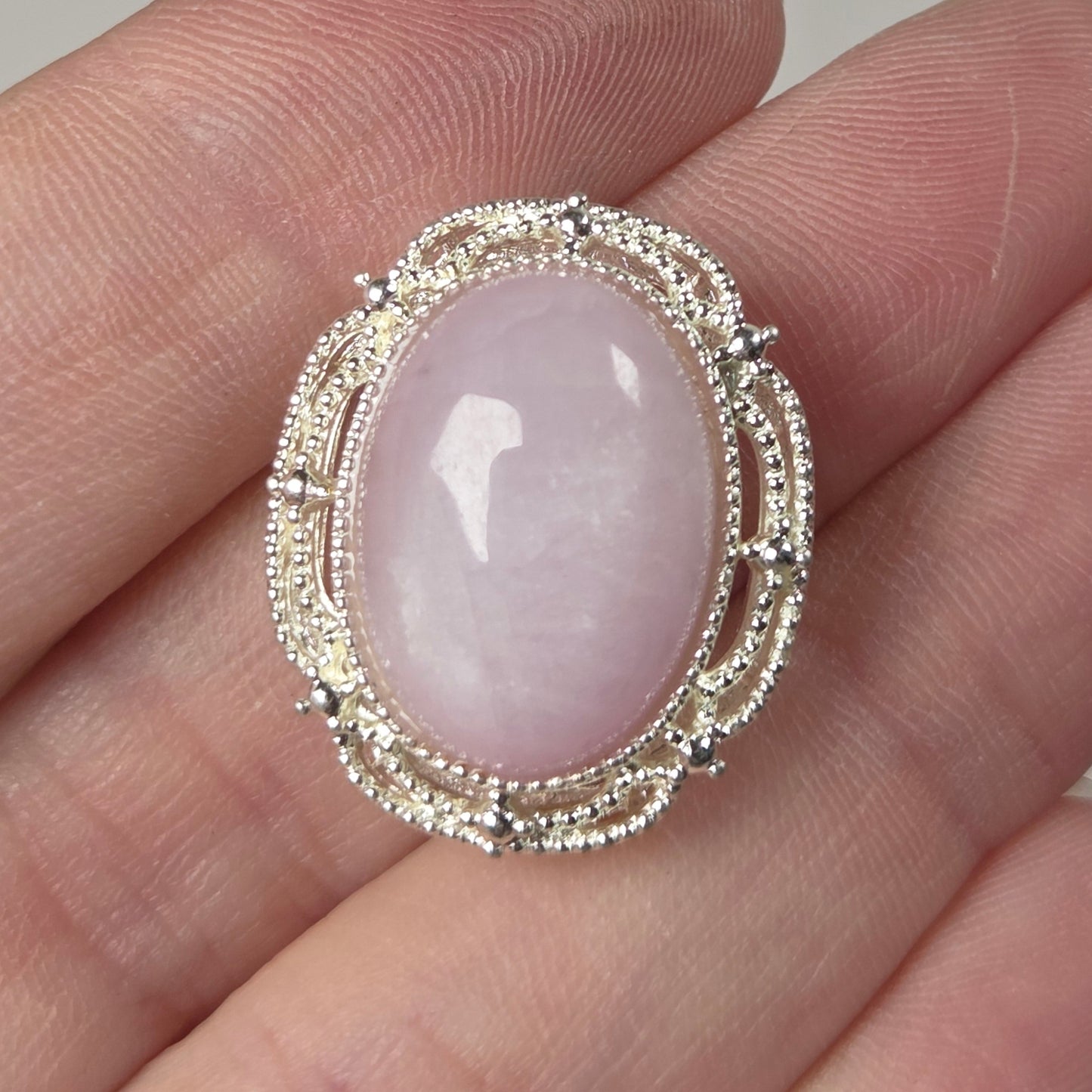 A silver-toned ring with an oval pale pink stone, held on a person's fingers against a plain light background.