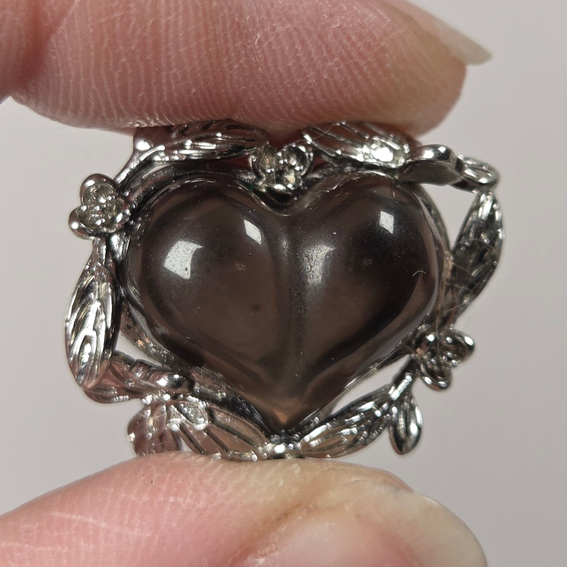 A silver ring with a heart-shaped dark smoky gemstone, held between two fingers against a plain light background.