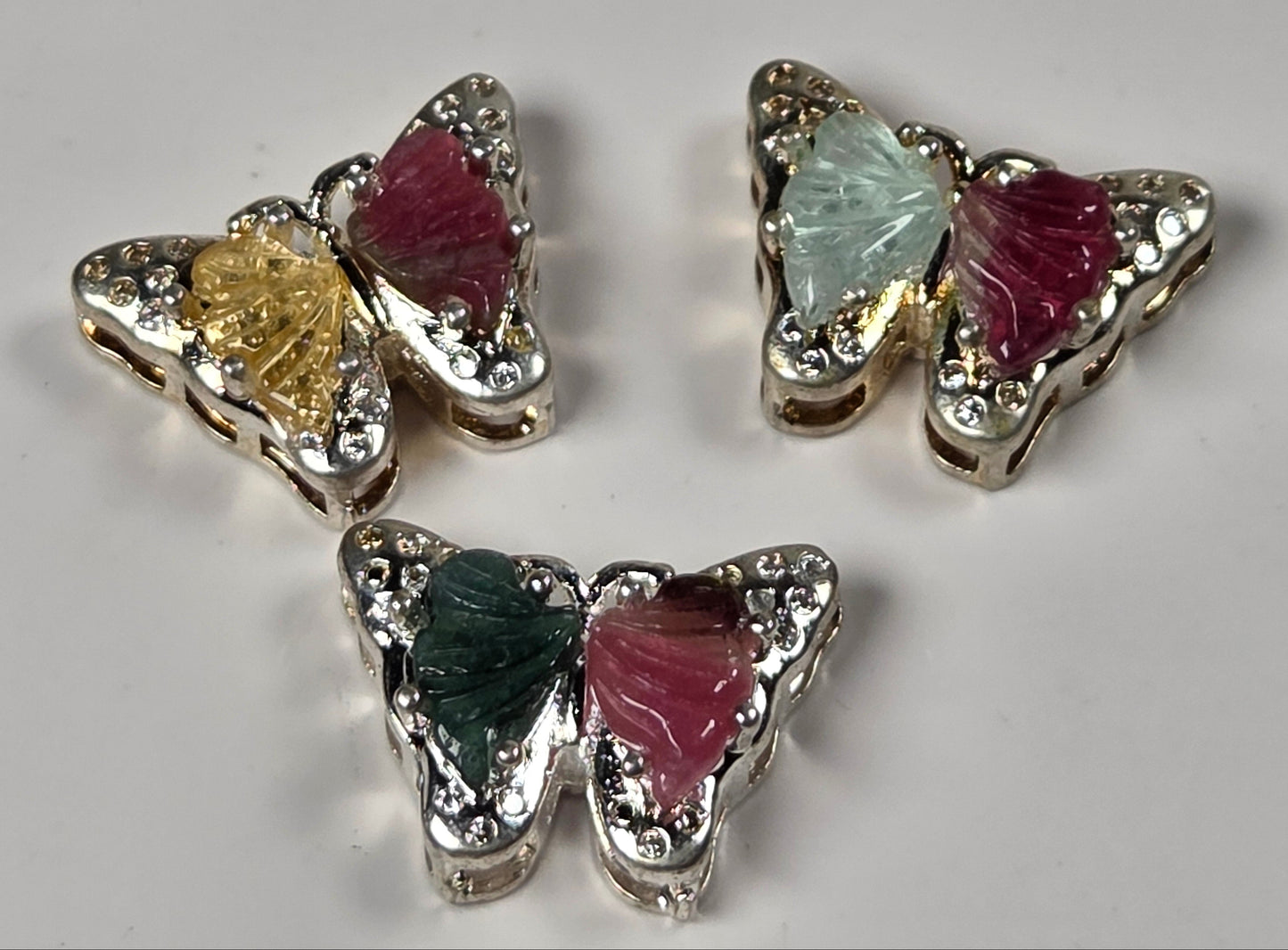 Three butterfly-shaped earrings with silver-toned metal borders, clear rhinestones, and colorful inlay wings in green, yellow, pink, and light blue, arranged on a light gray background.