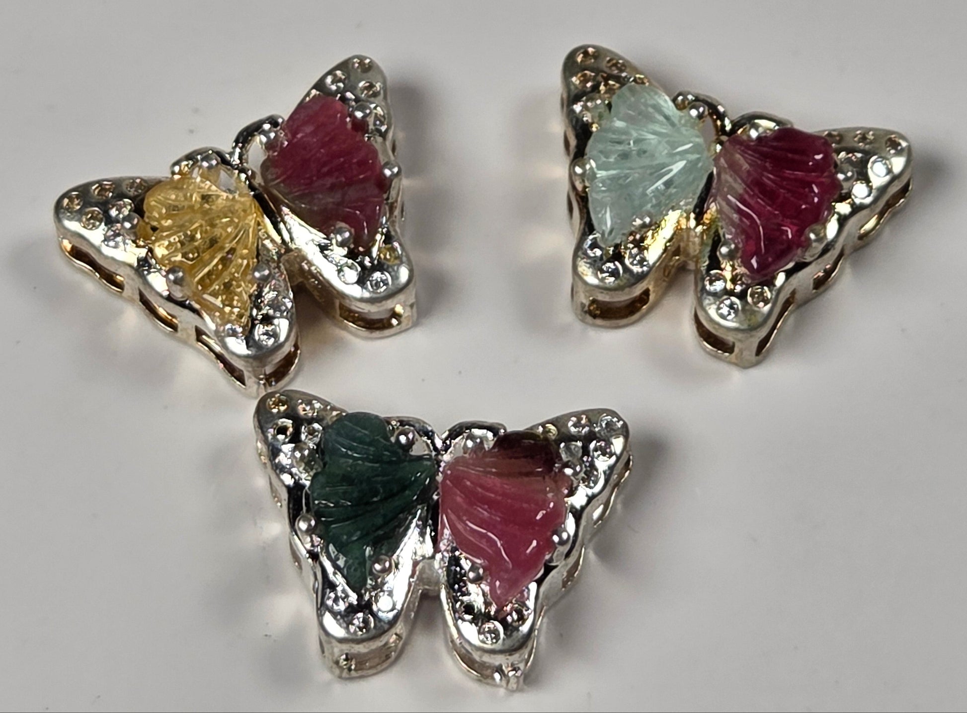 Three butterfly-shaped earrings with silver-toned metal borders, clear rhinestones, and colorful inlay wings in green, yellow, pink, and light blue, arranged on a light gray background.