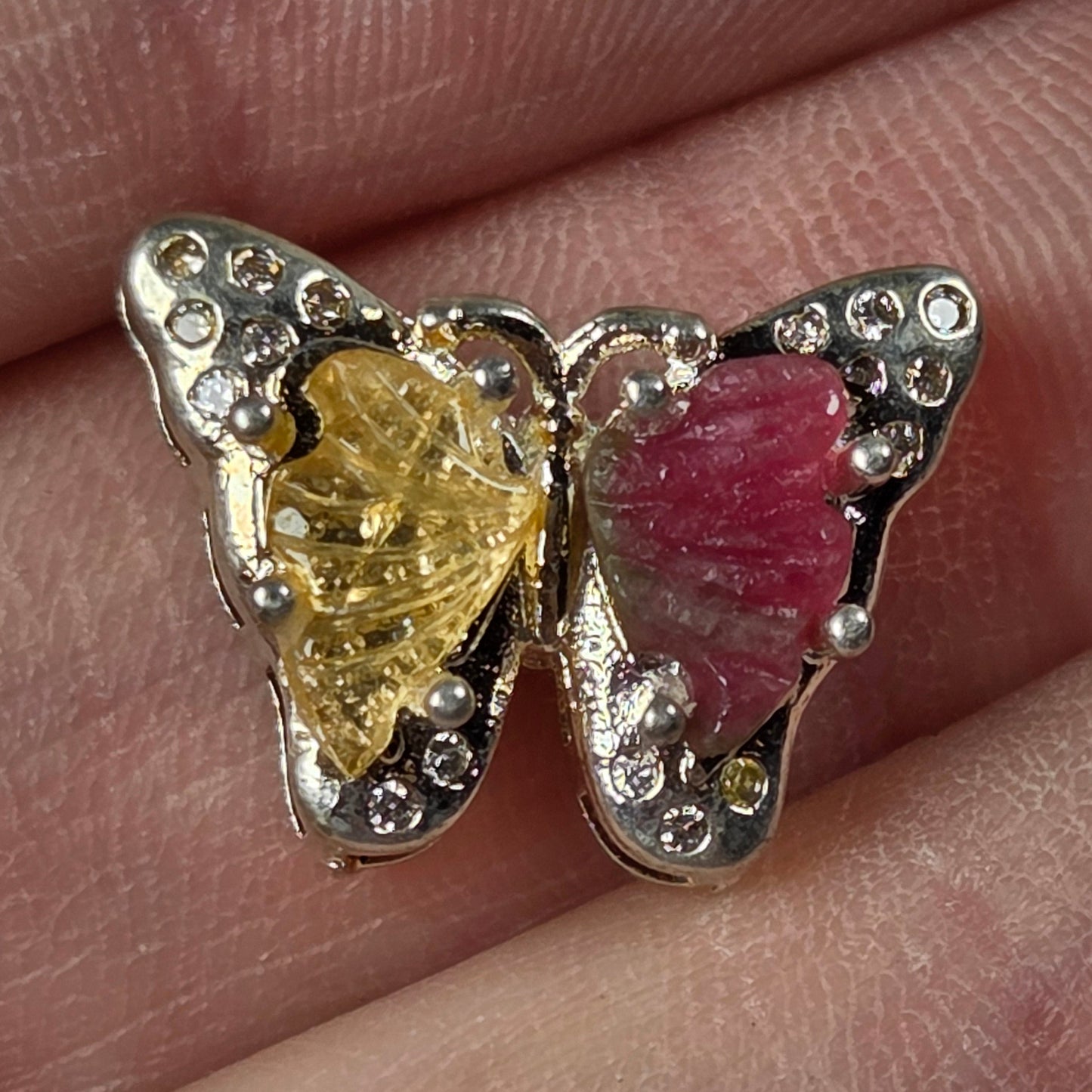 A butterfly-shaped ring with one pink wing and one yellow wing, set in silver with small clear stones, shown worn on a finger.