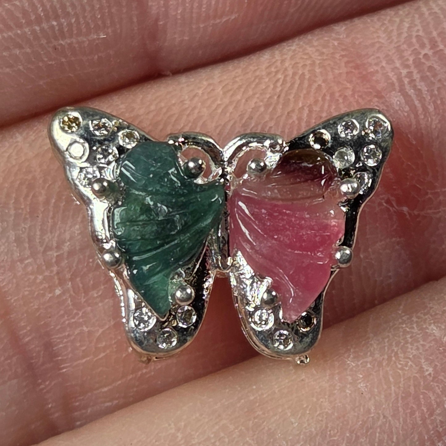 A butterfly-shaped ring with green and pink gemstone wings set in silver, resting on a person's fingers to show scale.
