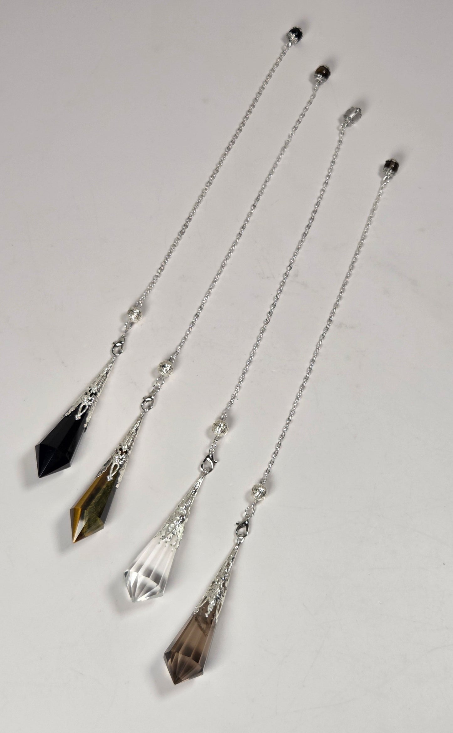 Four crystal and silver chain pendulums laid out diagonally on a light gray background. The pendulums feature faceted crystal drops in black, smoky brown, clear, and olive-brown tones, connected by delicate silver chains with small silver beads.
