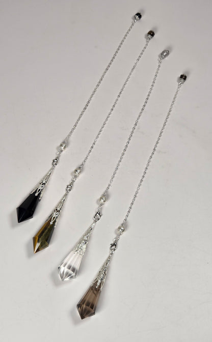 Four crystal and silver chain pendulums laid out diagonally on a light gray background. The pendulums feature faceted crystal drops in black, smoky brown, clear, and olive-brown tones, connected by delicate silver chains with small silver beads.