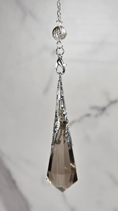 A silver-toned pendant pendulum  featuring a faceted smoky quartz crystal, displayed on a light gray marble background.
