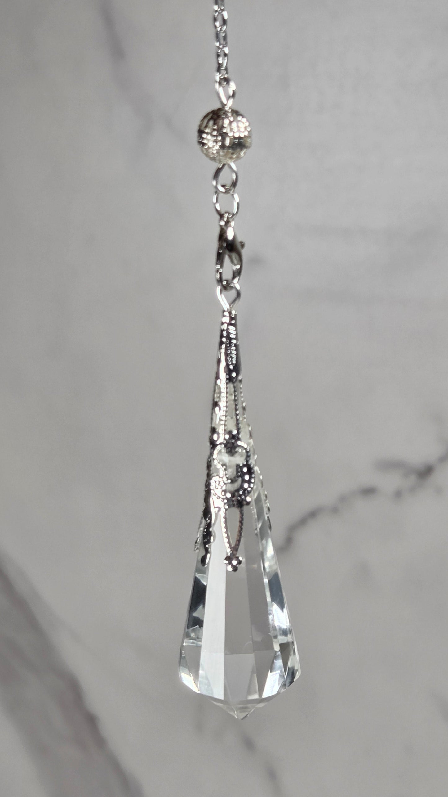 A silver-toned chain accessory with a faceted clear crystal pendant, displayed horizontally against a light gray marble background.